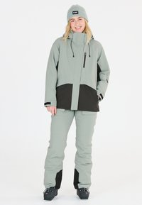 Kvinnors tvådelade vinteroutfit med en ljusgrön vattentät jacka med svarta detaljer och matchande ljusgröna byxor, båda med dragkedjefickor.