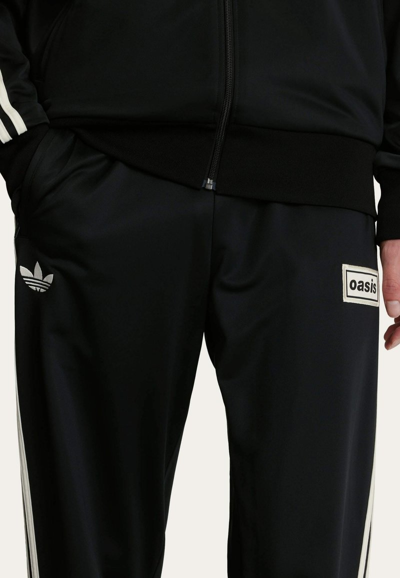 Sorte træningsbukser med hvide sidelister, prydet med et lille sort logo og et "oasis" mærke i en rektangulær hvid lap. Glat struktur.
