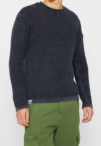 Marinblå stickad tröja med ett strukturerat mönster, rund halsringning och ribbade ärmslut. Har en liten logotagg vid nederkant.