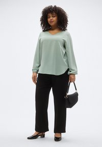 Ανοιχτό πράσινο μπλουζάκι με μακριά μανίκια και λαιμόκοψη σε σχήμα V, συνδυασμένο με μαύρο παντελόνι φαρδιάς γραμμής και μαύρα ψηλοτάκουνα από πατενταρισμένο δέρμα, κρατώντας μια μικρή μαύρη τσάντα.