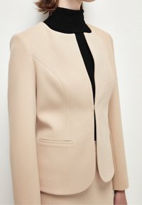 Beige kavaj med en strukturerad design, slät textur och rund hals. Har två framfickor och en lätt figursydd silhuett.