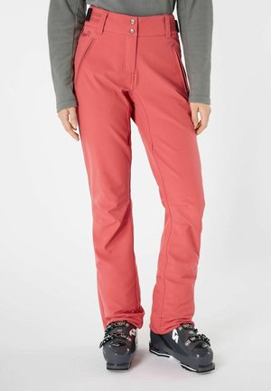 Femme portant un pantalon de neige corail et des chaussures de ski noires, debout sur un fond blanc uni.