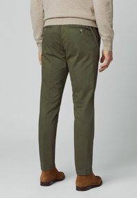 Homme portant un pantalon slim vert olive, un pull beige et des bottines en suède marron, debout devant un fond uni.