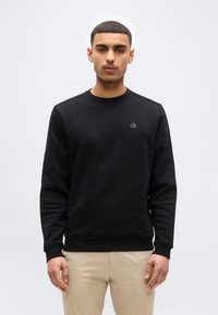 Νέος άνδρας που κοιτά μπροστά, φοράει μαύρο φούτερ Calvin Klein και μπεζ παντελόνι, πάνω σε απλό λευκό φόντο.