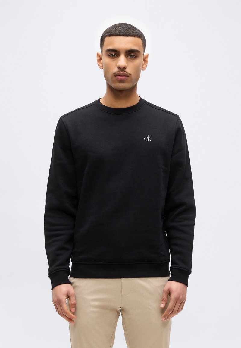 Νέος άνδρας που κοιτά μπροστά, φοράει μαύρο φούτερ Calvin Klein και μπεζ παντελόνι, πάνω σε απλό λευκό φόντο.