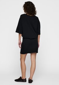 Vestido de algodón negro con mangas cortas, ajustado en la cintura y ligeramente acampanado en el dobladillo. Modelo con zapatos oscuros, visto desde la parte trasera.