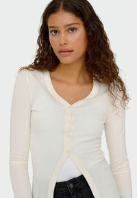 Cardigan a maniche lunghe a coste in bianco sporco, con scollo a V, chiusura con sette bottoni e design aperto sul davanti. Realizzato in un tessuto morbido e leggero.