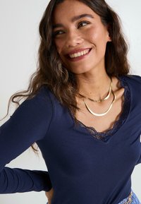 Top de manga larga en color navy con un borde de encaje en el escote, combinado con collares dorados en capas. Tejido cálido y texturizado; ajuste suave en el cuerpo.