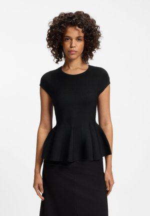 Femme aux cheveux bouclés portant un top noir cintré à basque et une jupe noire, debout devant un fond clair uni.