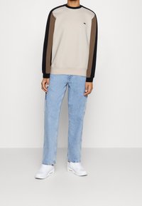 Persoon die een beige sweatshirt draagt met zwarte en bruine mouwen, lichtblauwe rechte jeans en witte sneakers op een effen achtergrond.