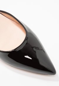 Chaussure à talon aiguille en cuir verni noir à bout pointu avec un intérieur rose pâle sur fond blanc.