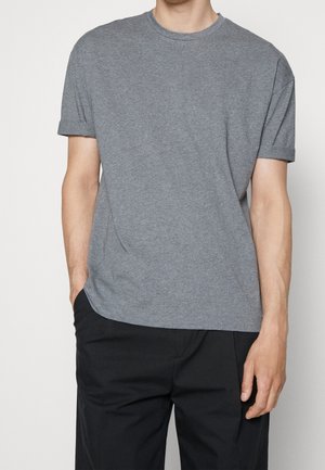 Mann in einem schlichten grauen T-Shirt mit kurzen Ärmeln und hochgerollten Ärmeln, trägt schwarze Hosen und steht mit einer Hand in der Tasche.