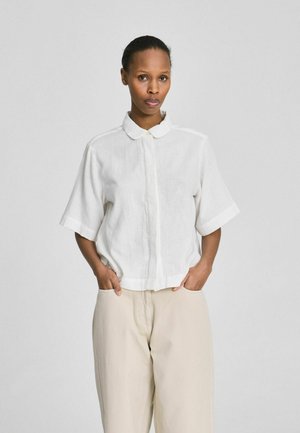 Vrouw met handen in de zakken, draagt een loszittend wit overhemd met korte mouwen en beige broek met wijde pijpen, tegen een effen lichte achtergrond.