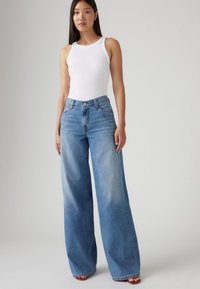 Jeans de denim azul de pierna ancha con un lavado descolorido, cintura alta y bolsillos delanteros, combinados con una camiseta blanca ajustada.