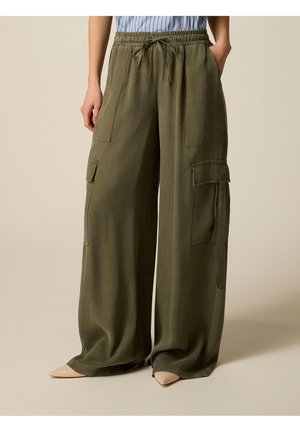 Vrouw draagt losse olijfgroene cargobroek met grote zakken en beige puntige schoenen, staand met handen in de zakken.