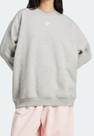 Femme portant un sweat-shirt Adidas gris clair avec un logo sur la poitrine et un pantalon ample rose clair, debout, les mains dans le dos.