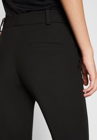 Pantalons noirs ajustés avec une texture lisse, dotés d'une poche arrière et d'un design slim, présentés de la taille vers le bas avec une main placée près du côté.