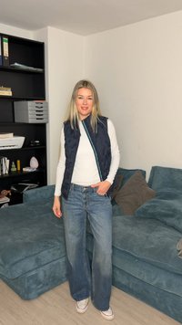 Γυναίκα με μακριά ξανθά μαλλιά, που φοράει λευκό πουκάμισο με μακριά μανίκια, μπλε γιλέκο, φαρδιά τζιν και λευκά αθλητικά παπούτσια, στέκεται στο καθιστικό κοντά σε μπλε καναπέ.