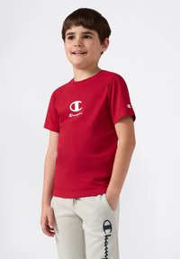 Junge mit braunen Haaren, der ein rotes Champion-T-Shirt und hellgraue Champion-Sweatpants trägt, mit einer Hand in der Tasche steht und lächelt.