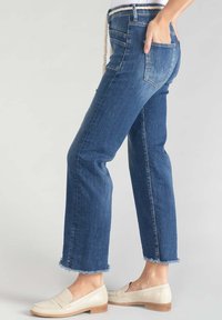 Jean en denim bleu foncé avec une jambe droite, un ourlet effiloché et des poches arrière, associés à des mocassins de couleur claire et une ceinture en corde.