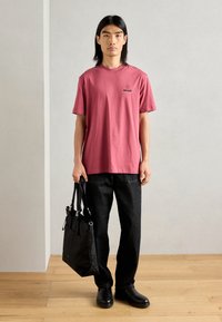 Un tricou din bumbac roz cu guler rotund și un logo negru, asortat cu pantaloni din denim închis și pantofi din piele negri, ținând o geantă tote neagră.