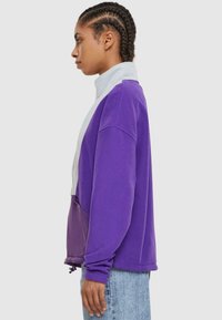 Lilla fleece genser med grå høy krage, med en glidelåslomme og elastisk snor i kanten, sammen med lyseblå jeans.