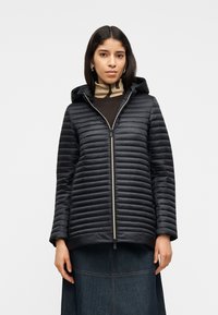 Γυναίκα που φοράει μαύρο καπιτονέ μπουφάν με κουκούλα πάνω από καφέ ζιβάγκο και σκουρόχρωμη τζιν φούστα, στέκεται μπροστά σε λευκό απλό φόντο.