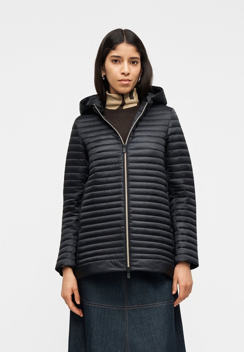 Γυναίκα που φοράει μαύρο καπιτονέ μπουφάν με κουκούλα πάνω από καφέ ζιβάγκο και σκουρόχρωμη τζιν φούστα, στέκεται μπροστά σε λευκό απλό φόντο.