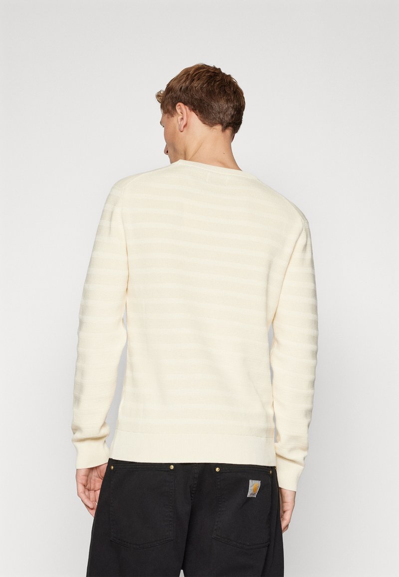 Mężczyzna z krótkimi, brązowymi włosami w kremowym, paskowanym swetrze z długim rękawem oraz czarnych spodniach, odwrócony plecami na tle jednolitego białego tła.