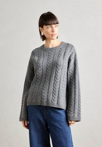 Pull gris en maille torsadée avec un col rond, des poignets et un ourlet côtelés. La texture présente des motifs en relief, associé à un jean en denim bleu.