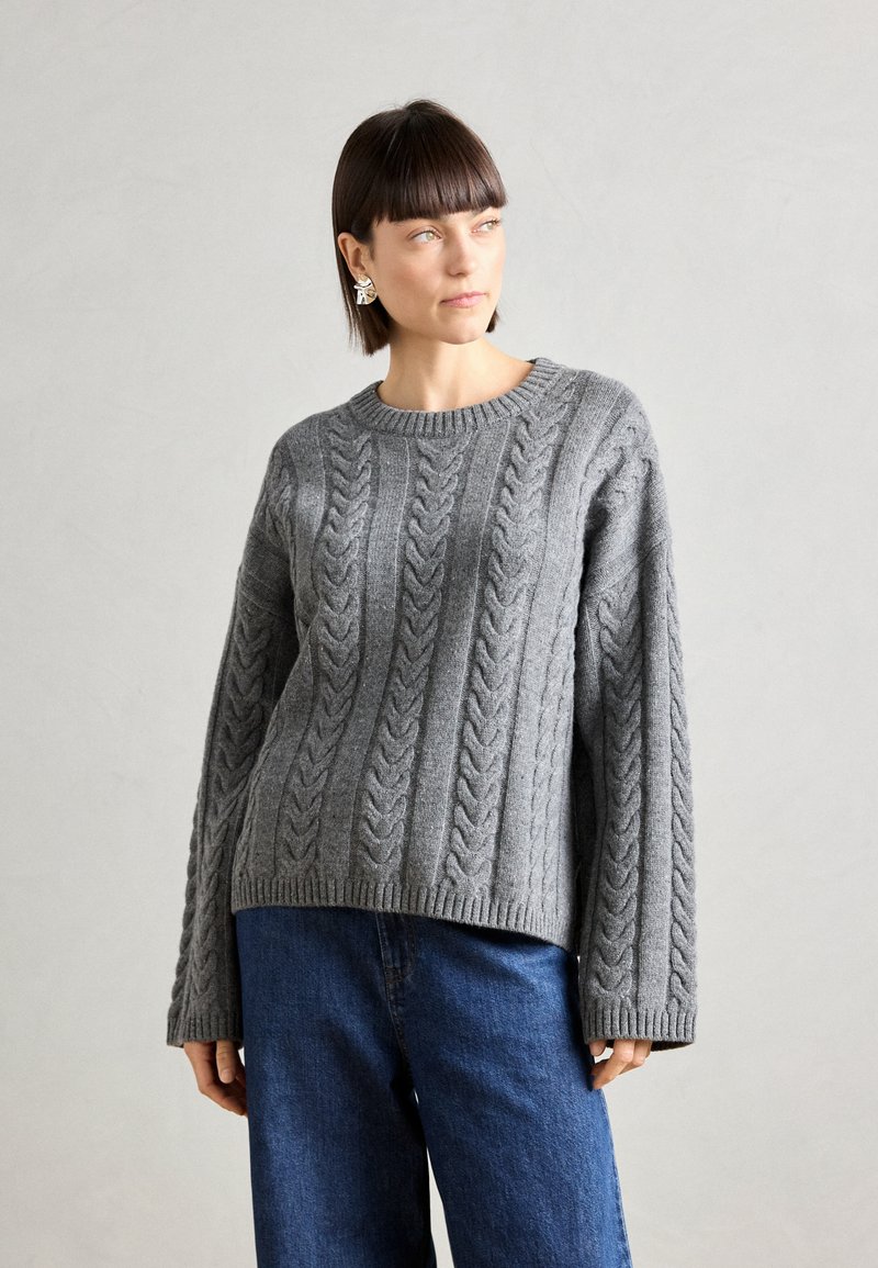 Pull gris en maille torsadée avec un col rond, des poignets et un ourlet côtelés. La texture présente des motifs en relief, associé à un jean en denim bleu.