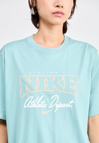 Világoskék pamut póló, élénk narancssárga "NIKE" logóval, a "Just Do It" felirattal a felett és az "Athletic Depart." szöveggel az alatt. Rövid ujjak.