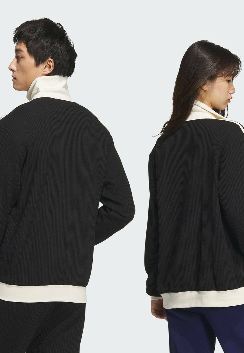 Chaqueta de tela de canalé negra con cuello alto, que presenta un acento blanco en el cuello y mangas texturizadas. Diseño unisex mostrado desde atrás.