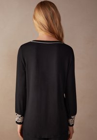Schwarzes Langarmshirt mit zarten weißen Spitzenakzenten an den Ärmelbündchen und einem dezenten weißen Saum am Ausschnitt. Glatte Stofftextur.
