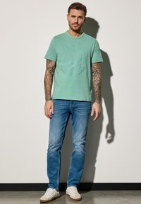 Muž s tetováním na celém rukávu stojí oblečený v tričku s tenisovým motivem v mentolové barvě, modrých džínách a bílých teniskách před prázdnou zdí.