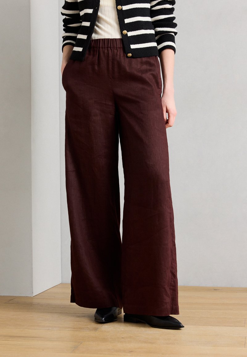 Woman wearing high-waisted, wide-leg dark brown pants, black pointed shoes, white ribbed top, and black-and-white striped cardigan with gold buttons.