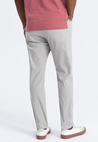 Chinos gris clair, coupe slim, en tissu coton. Dotés de deux poches arrière avec des coutures discrètes, portés avec des baskets blanches et une chemise rouge.