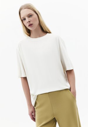 Ung kvinde med langt blondt hår iført en løs hvid kortærmet top og sennepsfarvede bukser, står foran en ensfarvet hvid baggrund.