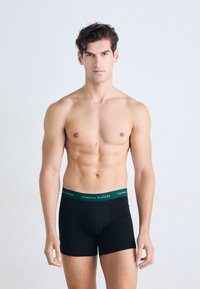 Shirtless man with short dark hair wearing black Tommy Hilfiger boxer briefs against a plain light background.