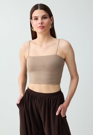 Jonge vrouw met stijl bruin haar, die gouden oorringen draagt, een beige cropped tanktop en losse donkerbruine broek met handen in de zakken.