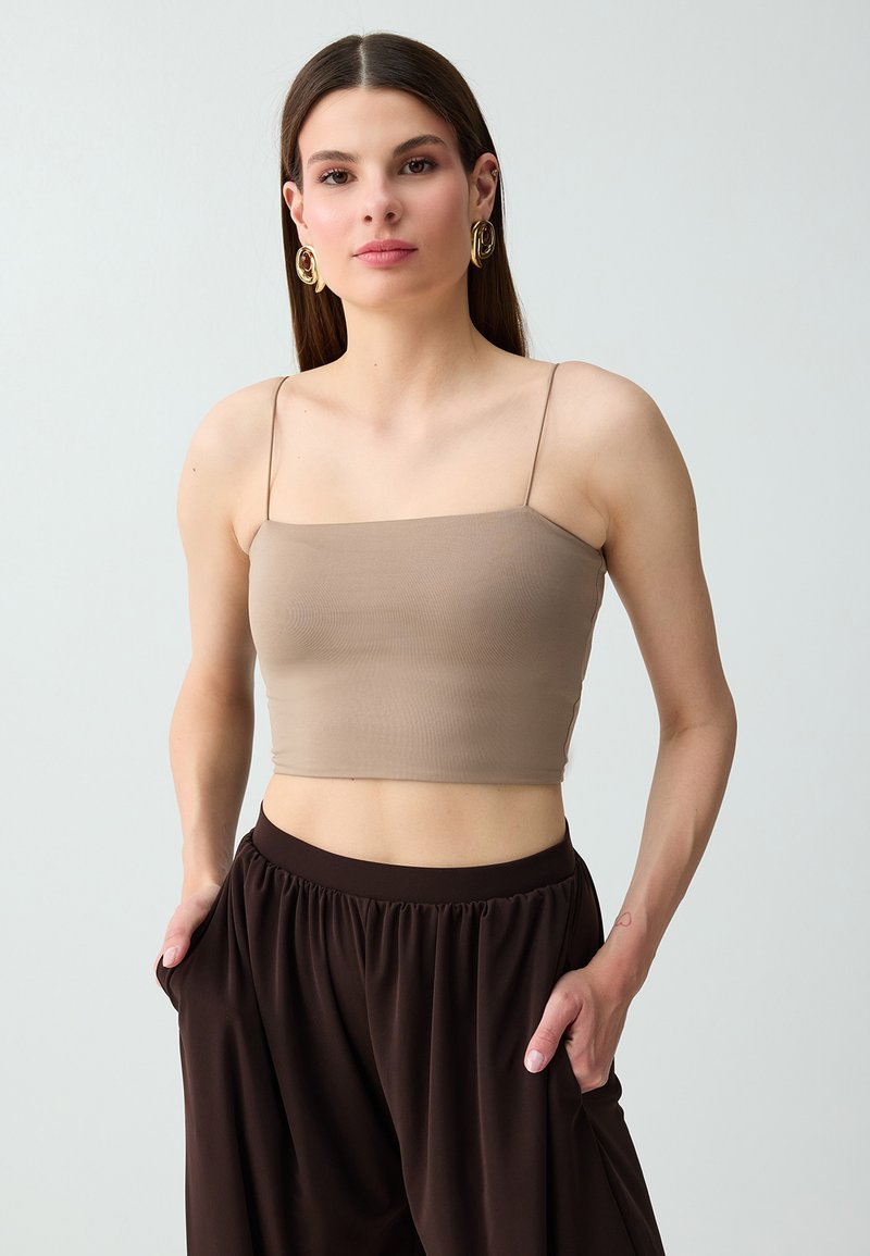 Jeune femme aux cheveux bruns et raides, portant des boucles d'oreilles créoles dorées, un débardeur court beige et un pantalon ample marron foncé avec les mains dans les poches.