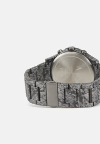 Montre grise avec un bracelet texturé présentant un motif marbré, des éléments en bronze et un boîtier rond métallique avec un dos réfléchissant.