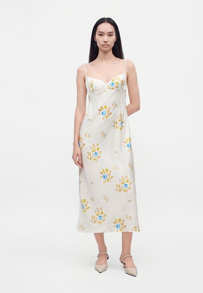 Femme aux longs cheveux foncés portant une robe midi blanche sans manches avec des motifs floraux bleus et jaunes et des chaussures pointues beiges.