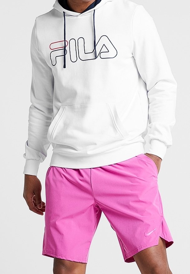 Homme portant un sweat à capuche blanc FILA et un short de sport Nike rose vif, debout avec une main dans la poche.