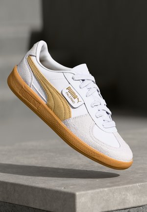 Weißer und beiger Puma Palermo Sneaker mit Gummisohle, in der Luft über einer Betonfläche vor dunkelgrauem Hintergrund gezeigt.