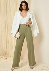Wijde olijfgroene broeken met hoge taille, gecombineerd met een witte tanktop en een oversized witte blazer. Het model houdt een crèmekleurige handtas vast.