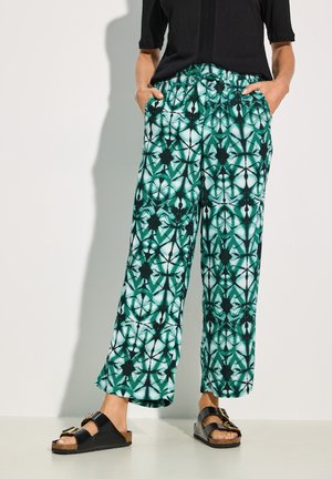 Groene gemusterde wijduitlopende broek met een elastische tailleband, voorzien van geometrische ontwerpen en pockets. G gedragen met zwarte sandalen.