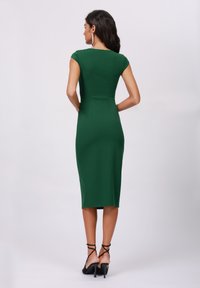 Grünes, tailliertes Midikleid mit kurzen Ärmeln und Schlitzen im Rücken. Glatte Textur mit einer eleganten Silhouette. Kombiniert mit schwarzen Riemchenschuhen.