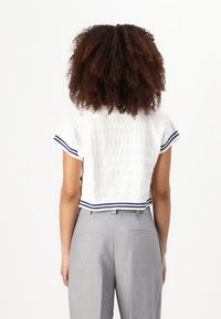 Top alb tricotat scurt, cu o țesătură texturată, având bordură albastră și albă cu dungi, asortat cu pantaloni gri deschis, croiți. Vedere din spate.