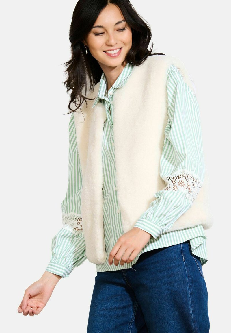 Vrouw lachend, gekleed in een witte nepbont vest over een groen-witte gestreepte blouse met kantuitsparingen op de mouwen, gecombineerd met blauwe jeans.
